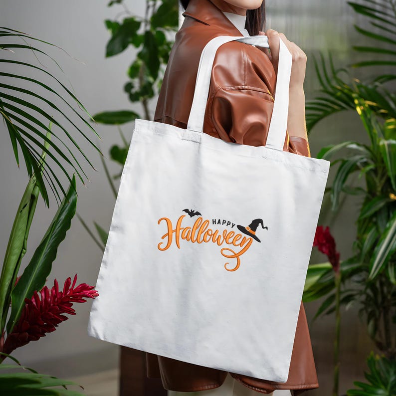 May include: A white tote bag with the words "Happy Halloween" in orange script, a black bat, and a witch's hat. The bag has white handles and is held by a person. The background features green plants and a brown jacket.