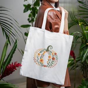 May include: White tote bag featuring a floral pumpkin design. The pumpkin is composed of orange, yellow, and green flowers and leaves, with a green stem. The bag has white handles and is held by a person.
