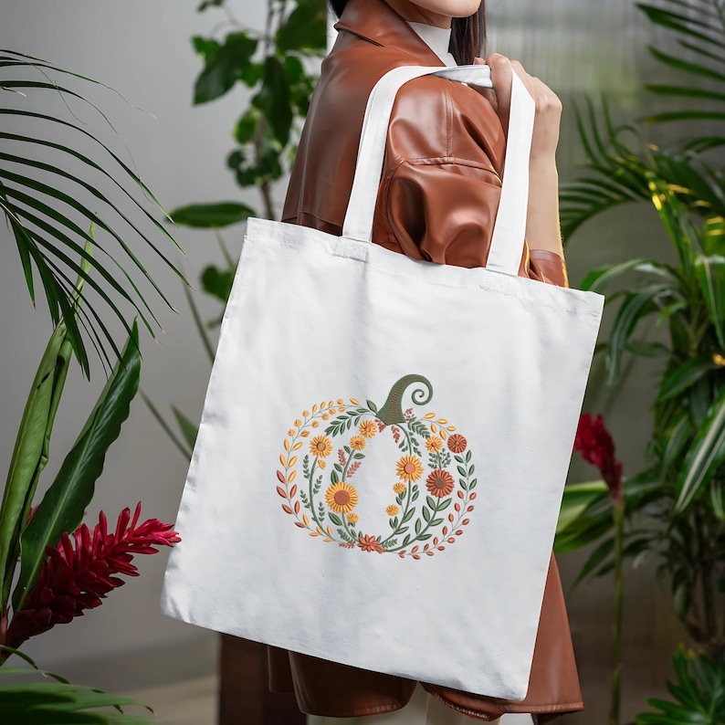 May include: White tote bag featuring a floral pumpkin design. The pumpkin is composed of orange, yellow, and green flowers and leaves, with a green stem. The bag has white handles and is held by a person.