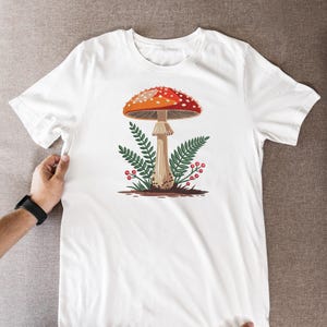 May include: White t-shirt featuring a colorful illustration of a mushroom with a red and white spotted cap, brown stem, and green foliage. The design is centered on the shirt, creating a nature-inspired aesthetic.