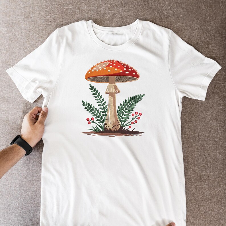 May include: White t-shirt featuring a colorful illustration of a mushroom with a red and white spotted cap, brown stem, and green foliage. The design is centered on the shirt, creating a nature-inspired aesthetic.