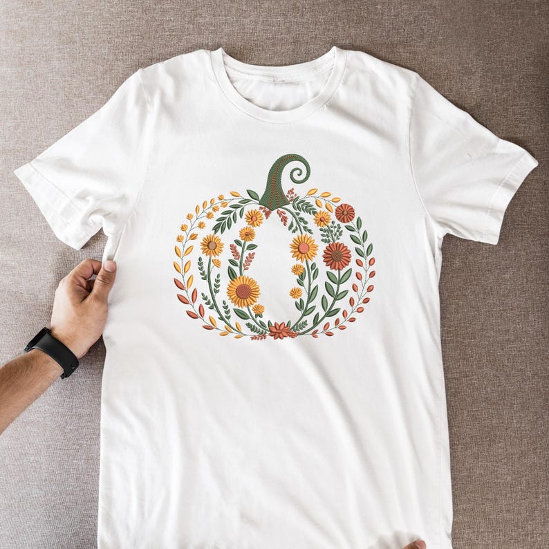 May include: White t-shirt featuring a floral pumpkin design. The pumpkin is composed of orange, yellow, and red flowers, green leaves, and a green stem. The design is centered on the front of the shirt, creating a fall or autumn theme.