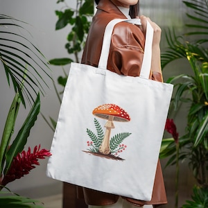 May include: White tote bag featuring a colorful illustration of a mushroom with a red and white spotted cap, a beige stem, and green foliage. The bag has white handles and is made of a sturdy fabric, perfect for carrying groceries or books.
