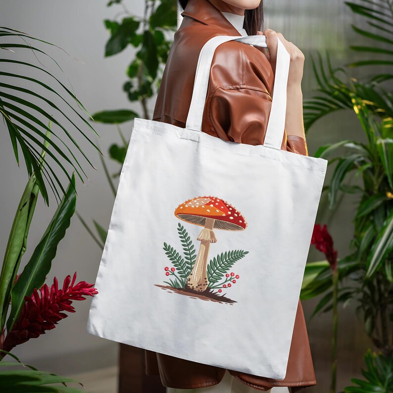 May include: White tote bag featuring a colorful illustration of a mushroom with a red and white spotted cap, a beige stem, and green foliage. The bag has white handles and is made of a sturdy fabric, perfect for carrying groceries or books.