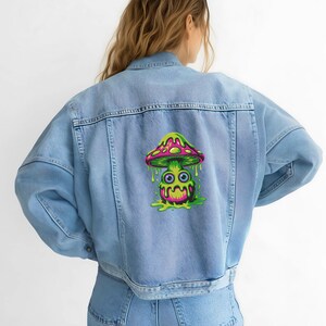 Op de afbeelding: Lichtblauwe spijkerjas met een kleurrijke paddenstoelafbeelding op de rug. De paddenstoel heeft een roze en groene hoed, een groene basis en een cartoongezicht met druipende details. De jas heeft een klassiek ontwerp.