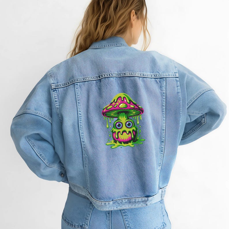 Op de afbeelding: Lichtblauwe spijkerjas met een kleurrijke paddenstoelafbeelding op de rug. De paddenstoel heeft een roze en groene hoed, een groene basis en een cartoongezicht met druipende details. De jas heeft een klassiek ontwerp.