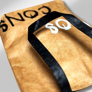 Könnte beinhalten: Eine braune Papiertüte mit schwarzen Henkeln und weißem Schriftzug "SQ" auf der Vorderseite. Die Tasche hat eine strukturierte Oberfläche.