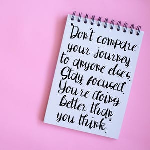 以下が含まれることがあります： ピンク色の背景に、黒い文字が書かれた白いリング綴じノート。テキストは「自分の旅を他の誰とも比較しないでください。集中してください。あなたは思っている以上にうまくいっています。」