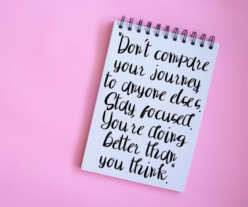 以下が含まれることがあります： ピンク色の背景に、黒い文字が書かれた白いリング綴じノート。テキストは「自分の旅を他の誰とも比較しないでください。集中してください。あなたは思っている以上にうまくいっています。」