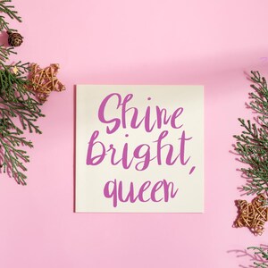 以下が含まれることがあります： 紫色の筆記体で「Shine bright, queen」と書かれた正方形のクリーム色のカード。カードはピンク色の背景にあり、常緑樹の枝、金の星の飾り、松ぼっくりで飾られています。