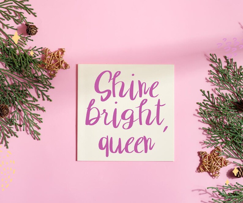 以下が含まれることがあります： 紫色の筆記体で「Shine bright, queen」と書かれた正方形のクリーム色のカード。カードはピンク色の背景にあり、常緑樹の枝、金の星の飾り、松ぼっくりで飾られています。