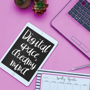 以下が含まれることがあります： ピンク色のノートパソコン、タブレット、ウィークリーアジェンダがピンク色の表面に配置されています。タブレットには、白いスクリプトで「Digital space dreamy mind」という言葉が表示されています。アジェンダは、黒と白のストライプで「Weekly Agenda」という言葉が書かれています。小さな鉢植えの植物も表示されています。