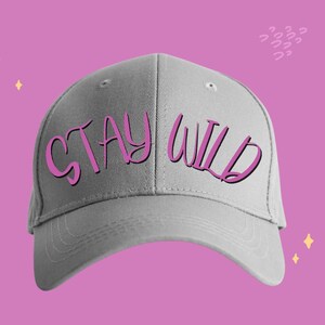 以下が含まれることがあります： 前面にピンク色の「STAY WILD」の文字が入った、ライトグレーの野球帽。装飾的な要素のあるピンク色の背景に帽子が置かれています。
