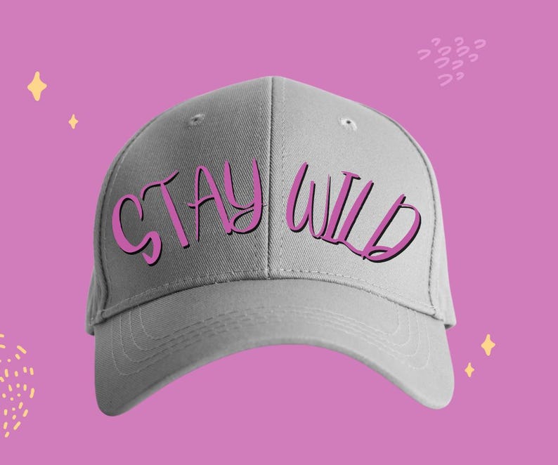 以下が含まれることがあります： 前面にピンク色の「STAY WILD」の文字が入った、ライトグレーの野球帽。装飾的な要素のあるピンク色の背景に帽子が置かれています。