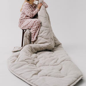 May include: A light beige quilted blanket and matching pillow are draped over a wooden stool. The blanket is on the floor. The person is wearing a pink and white checked pajama set.