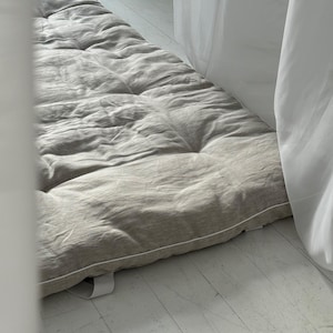 Op de afbeelding: Een neutraal getuft vloerkussen of matras. Het rechthoekige kussen is lichtgrijs met een witte bies en een witte band. Het kussen ligt op een witte houten vloer, met witte gordijnen op de achtergrond.