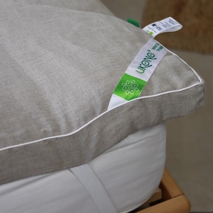 Op de afbeelding: Close-up van een lichtgrijze matrasbeschermer met een witte rand en een groen-wit label met de tekst "LIKONO SINCE 1999". De topper ligt op een wit matras met een witte elastische band.