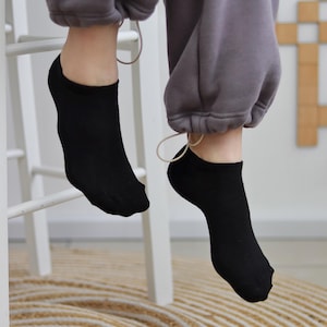May include: A close-up shot of two black no-show socks. The socks are worn with gray sweatpants. The person is sitting on a white chair, and the floor has a round, woven rug.