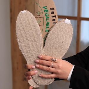 Puede incluir: Plantillas beige de forma ovalada hechas de un material natural, sujetas con una etiqueta de cartón. La etiqueta dice "Vegetable Insole" y declara "100% natural material". Las plantillas parecen estar hechas de tela tejida.