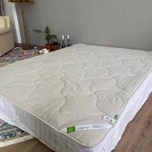 May include: A cream mattress topper with a quilted design, placed on a bed. The topper has rounded edges and a tag with the text "UKONO". The bed is in a room with a wooden floor and a grey wall.