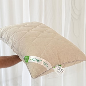 Op de afbeelding: Een beige rechthoekig kussen met een gewatteerd patroon. Het kussen wordt tegen een wit gordijn gehouden. Een label met groene tekst en de merknaam "UKONO" is bevestigd.