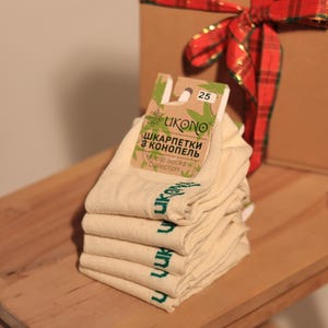 Könnte beinhalten: Ein Stapel beiger Hanfsocken mit grünem Text, beschriftet mit "UKONO Hemp Socks Collection", liegt auf einer Holzoberfläche. Im Hintergrund befindet sich eine Geschenkbox mit einem rot-grünen Karoband. Die Socken haben ein Etikett mit der Nummer 25.
