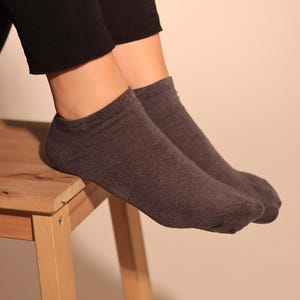 Puede incluir: Un par de calcetines grises oscuros sin costuras. Los calcetines están hechos de un material de punto suave. La persona lleva leggings negros. Los calcetines descansan sobre un pequeño taburete de madera.