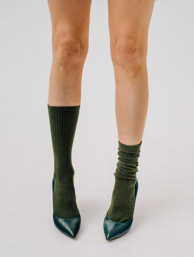 May include: A pair of legs wearing dark green socks and teal pointed-toe high heels. The socks are a solid color and have a ribbed texture. The heels are a glossy teal color.
