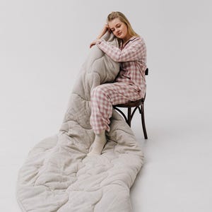 May include: A light beige quilted blanket with a diamond pattern is draped over a wooden chair. The blanket is paired with a matching beige pillow. The person in the image is wearing pink and white plaid pajamas.