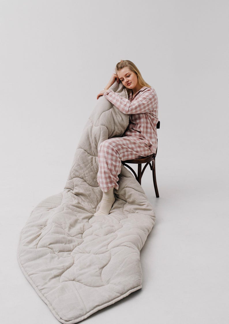 May include: A light beige quilted blanket with a diamond pattern is draped over a wooden chair. The blanket is paired with a matching beige pillow. The person in the image is wearing pink and white plaid pajamas.
