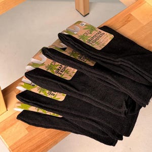 May include: A stack of black socks with attached cardboard labels. The labels have green and brown accents and text in a foreign language. The socks are neatly arranged on a wooden surface.