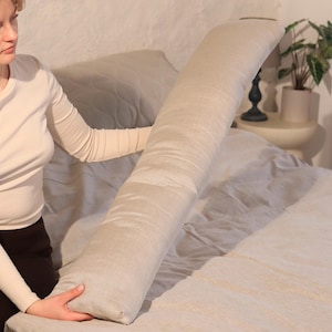 Op de afbeelding: Een lang, beige lichaamskussen ligt op een bed met bijpassend beddengoed. Het kussen is cilindrisch en lijkt gemaakt van een zachte, getextureerde stof. Het bed staat in een kamer met een neutraal kleurenpalet.