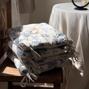 May include: A stack of four square seat cushions with a floral pattern in shades of blue and white. The cushions are tied together with twine and have fabric ties on the corners. They are placed on a wooden chair.