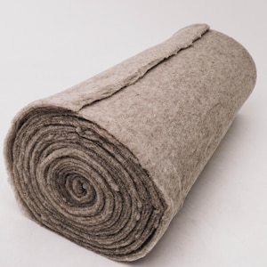 May include: A rolled-up, thick, beige fabric. The material appears to be a dense, felt-like textile, with a slightly uneven texture. The roll is tightly wound, revealing the layers of the fabric. The edges are slightly frayed.