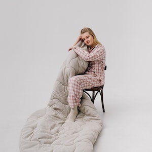 May include: A person wearing pink and white plaid pajamas sits on a wooden chair, with a large, light gray quilted blanket draped over the chair and the floor. The blanket has a textured, wavy pattern.