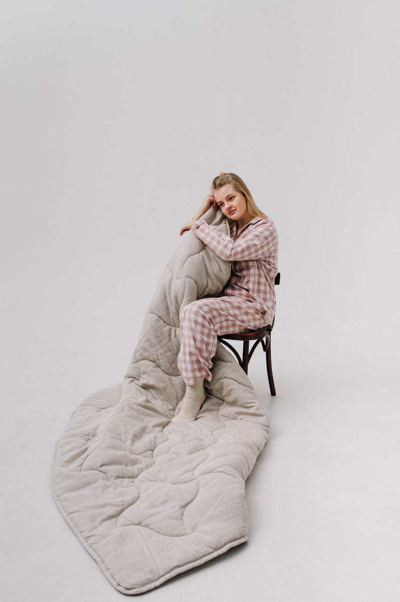 May include: A person wearing pink and white plaid pajamas sits on a wooden chair, with a large, light gray quilted blanket draped over the chair and the floor. The blanket has a textured, wavy pattern.