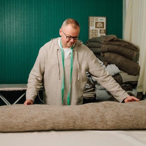 May include: A man in a light brown hoodie and glasses is working with a large, rolled-up, tan fabric on a white surface. A green measuring tape hangs around his neck. Behind him is a stack of similar fabrics and a teal wall.