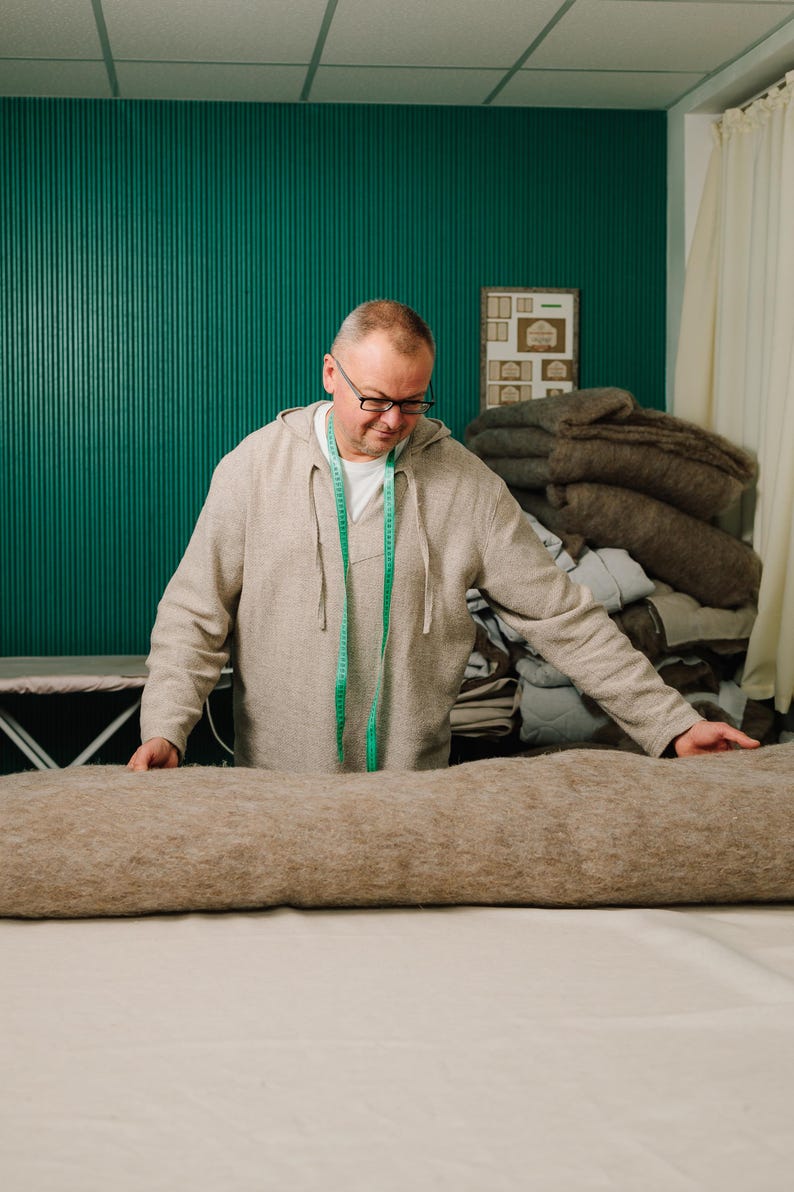 May include: A man in a light brown hoodie and glasses is working with a large, rolled-up, tan fabric on a white surface. A green measuring tape hangs around his neck. Behind him is a stack of similar fabrics and a teal wall.