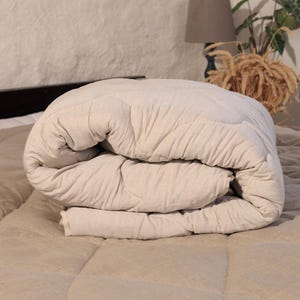 May include: A folded, beige-colored comforter on a bed. The comforter has a quilted pattern and is made of a soft, textured fabric. The bed is covered with a matching beige bedspread. The scene is lit by natural light, creating a warm and inviting atmosphere.