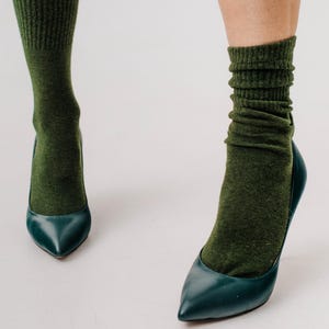 May include: A pair of teal-colored high heels with pointed toes. The shoes are paired with olive green socks, creating a contrast in textures and colors. The socks are slightly scrunched at the ankles.