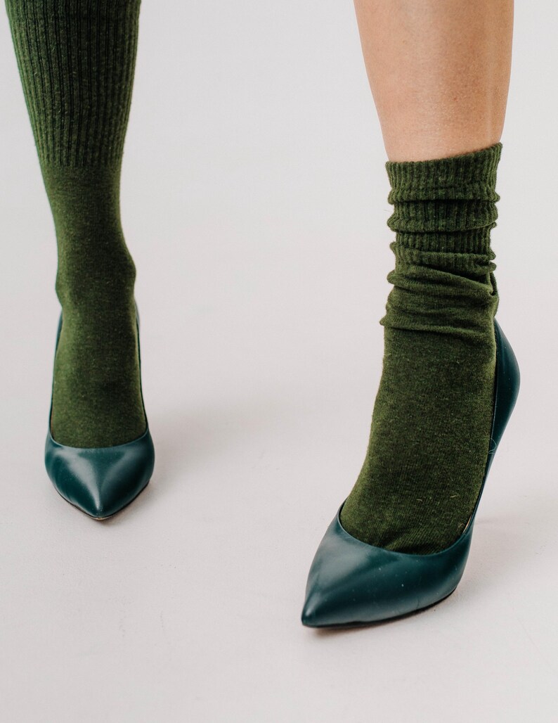 May include: A pair of teal-colored high heels with pointed toes. The shoes are paired with olive green socks, creating a contrast in textures and colors. The socks are slightly scrunched at the ankles.