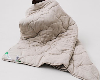 May include: A beige quilted throw blanket with a wavy pattern. The blanket is folded and draped over a chair.
