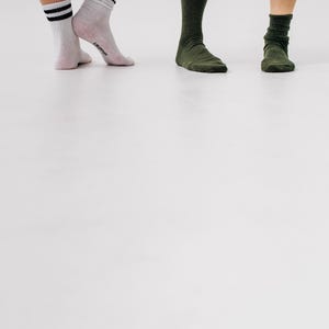 May include: Three pairs of socks in different colors. One pair is white with black stripes, another is light pink, and the last two pairs are dark green. The socks are worn by three people, with their legs and feet visible.