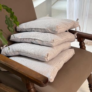 Op de afbeelding: Vier vierkante decoratieve kussens gestapeld op een bruine fauteuil. De kussens hebben een lichtgrijs geweven patroon met een decoratieve rand. De fauteuil heeft een bruine leren zitting en houten armen en poten.