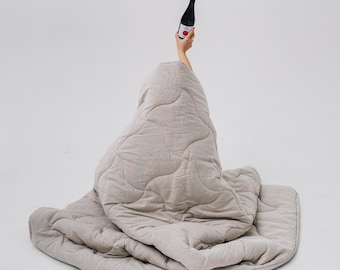 May include: A light grey quilted blanket with a person covered by it, holding a black wine bottle with a red label. The blanket has a wavy, stitched pattern. The background is plain white.