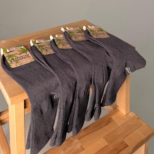 Könnte beinhalten: Ein Stapel grauer Socken mit Etiketten, präsentiert auf einem Holzschemel. Die Socken sind aus geripptem Material und haben ein kleines Logo. Die Etiketten zeigen Text in einer Fremdsprache.