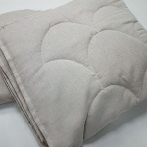 May include: Folded, light beige quilted blanket with a cloud-like pattern. The blanket is made of a textured fabric and appears soft. The edges are neatly finished, and the blanket is folded in a stack. The overall look is simple and cozy.