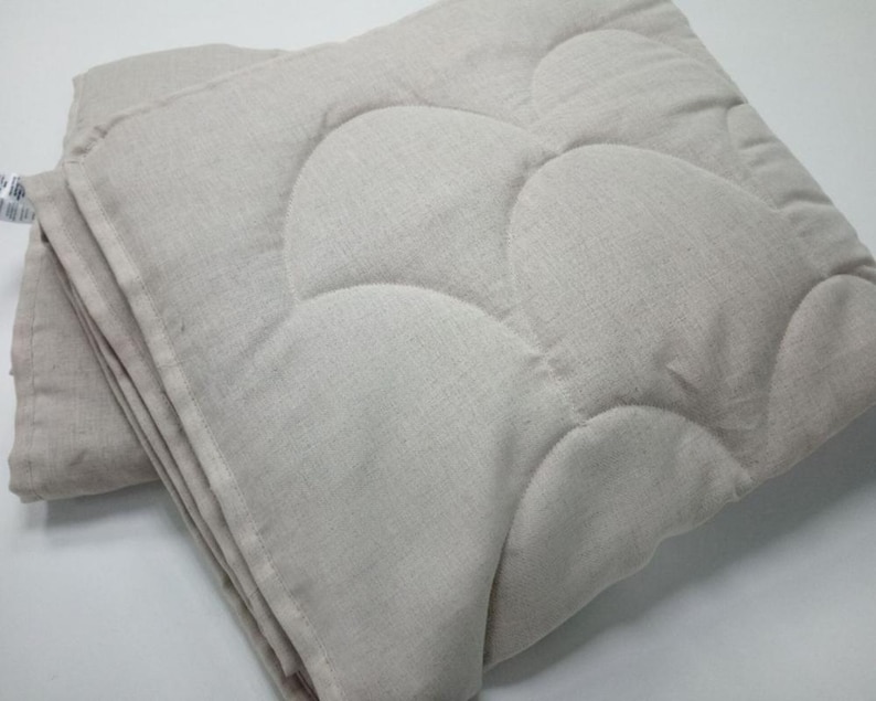 May include: Folded, light beige quilted blanket with a cloud-like pattern. The blanket is made of a textured fabric and appears soft. The edges are neatly finished, and the blanket is folded in a stack. The overall look is simple and cozy.