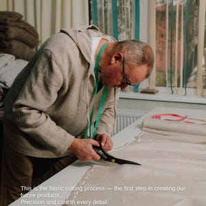 May include: A person cutting fabric with scissors on a white table. The fabric is beige, and a green measuring tape is around the person's neck. Text on the image reads: "This is the fabric cutting process - the first step in creating our future products. Precision and care in every detail."