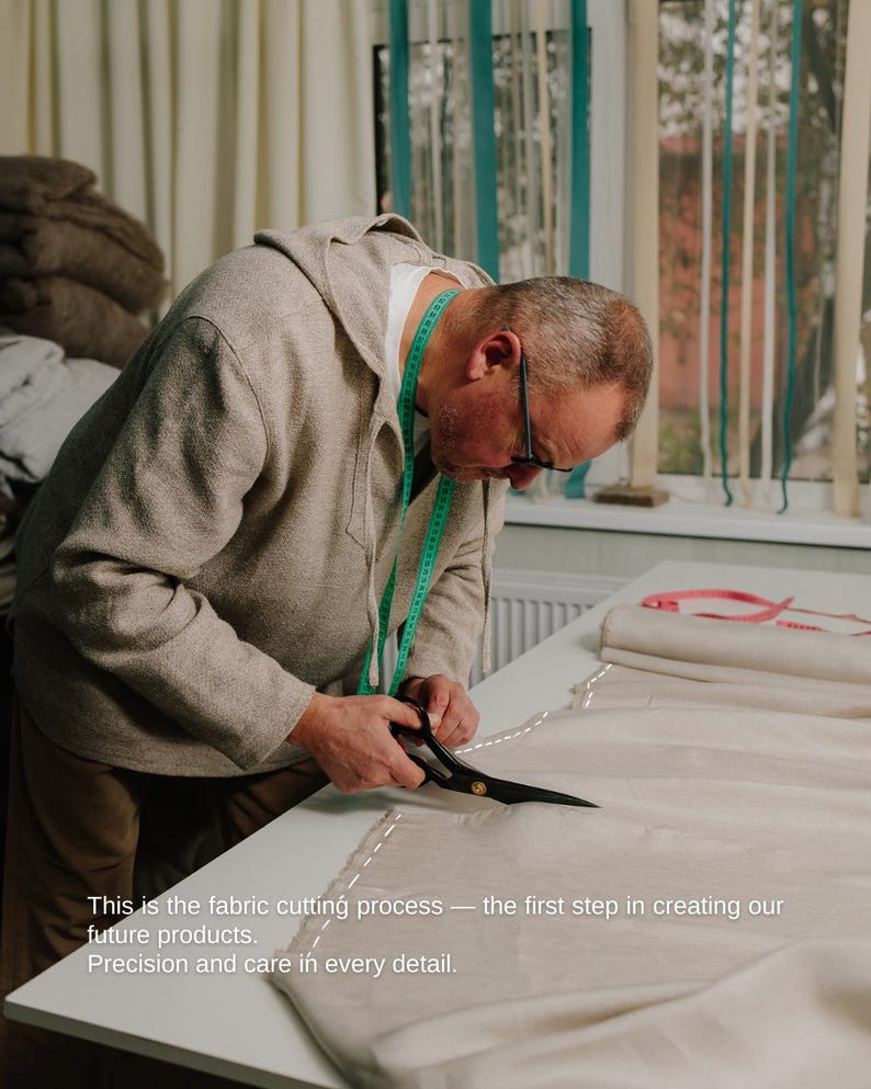 May include: A person cutting fabric with scissors on a white table. The fabric is beige, and a green measuring tape is around the person's neck. Text on the image reads: "This is the fabric cutting process - the first step in creating our future products. Precision and care in every detail."